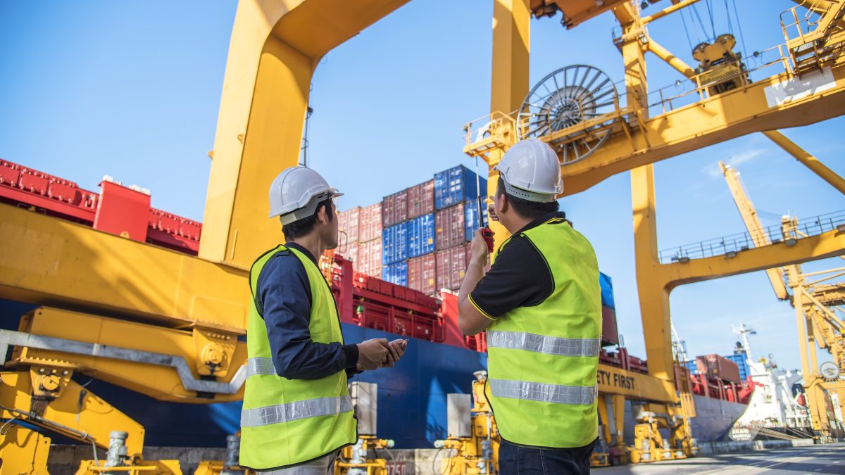 A picture of 2 workers in yellow vests on a port looking up at cargo ships and equipment