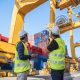 A picture of 2 workers in yellow vests on a port looking up at cargo ships and equipment