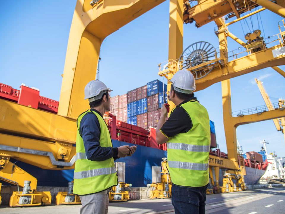 A picture of 2 workers in yellow vests on a port looking up at cargo ships and equipment